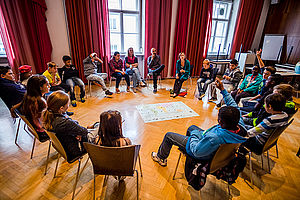 Sesselkreis in einem großen Saal. In der Mitte liegt eine Weltkarte