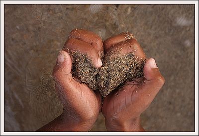 Zwei Hände formen ein Herz und halten dabei Sand. Im Hintergrund Meer und Sand