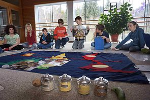 SchülerInnen beim Workshop "Was isst die Welt" im Sitzkreis. In ihrer Mitte verschiedene Materialien